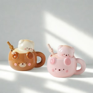 May include: Two ceramic mugs with bear-shaped lids and spoons. One mug is brown with a cream-coloured bear lid, the other is pink with a pink bear lid. Both mugs have a bear face design.
