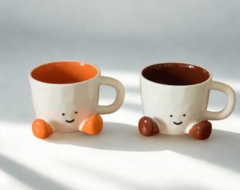 Peut inclure: Deux tasses en céramique au design fantaisiste. Les tasses sont blanches avec des intérieurs orange et marron. Chaque tasse a un visage souriant et des pieds orange ou marron. Les tasses ont une anse et sont posées sur un fond blanc.
