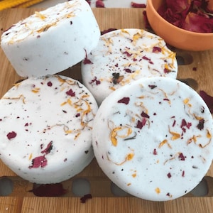 May include: Four white, round bath bombs with dried flower petals on a wooden surface. The bath bombs are decorated with red and yellow petals. Two small orange bowls are in the background, one with dried petals and the other with herbs.
