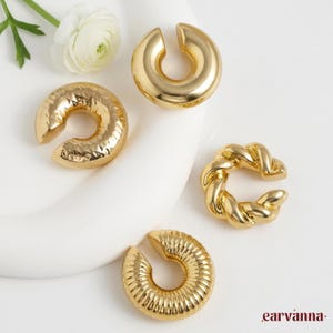 Puede incluir: Una colección de puños de oreja dorados en varios diseños. Los puños de oreja incluyen un diseño liso, martillado, de cuerda trenzada y acanalado. Se muestran sobre una superficie blanca con una flor blanca y hojas verdes.