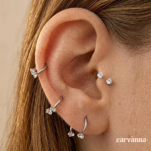 May include: Close-up of an ear adorned with multiple silver earrings. The earrings include small studs and hoop styles, some featuring clear, sparkling gemstones. The ear is set against a neutral background, highlighting the jewelry.