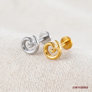 Könnte beinhalten: Zwei spiralförmige Ohrstecker, einer silberfarben und einer goldfarben, liegen auf einer strukturierten, cremefarbenen Oberfläche. Die Ohrringe haben einen flachen Rücken und ein geschwungenes, wirbelndes Design. Der goldene Ohrring hat eine glänzende, polierte Oberfläche.