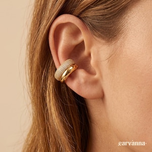 Könnte beinhalten: Ein goldener Ohrklemme mit einem Doppelband-Design. Das obere Band ist mit kleinen, funkelnden Steinen besetzt, während das untere Band glatt ist. Der Ohrklemme wird am Ohr getragen und unterstreicht seinen eleganten und modernen Stil.