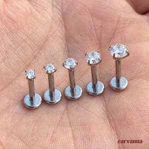 May include: Five silver-toned labret studs arranged in a row, each featuring a round, clear, faceted gemstone set in a four-prong setting. The posts are smooth and cylindrical, ending in a flat, circular base plate for secure placement.
