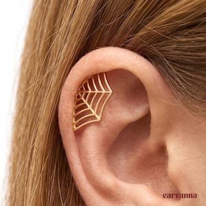 May include: A gold-toned ear cuff shaped like a spider web, featuring a geometric openwork design with radiating lines and concentric arcs. The piece is positioned along the upper cartilage of the ear, showcasing a polished metallic finish.