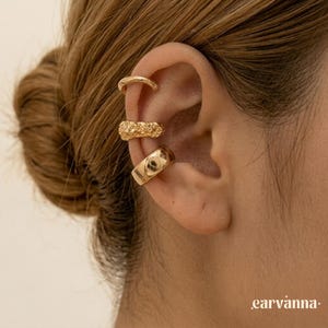 May include: Gold ear cuffs in various designs. One is a simple band, another has a textured, nugget-like appearance, and the third is a wider band with a subtle design. The ear cuffs are worn on the ear.