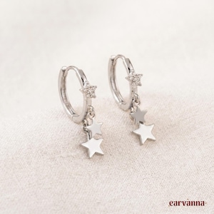 May include: Silver hoop earrings with star charms. Each earring has a small, sparkling star at the top and two white star charms hanging below. The earrings are shown on a light-coloured, textured surface.