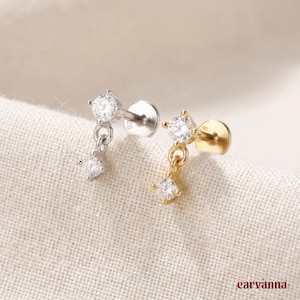 May include: Two stud earrings with dangling cubic zirconia stones. One earring is silver-toned, and the other is gold-toned. The earrings have a small, round stud that screws into the ear. The dangling stones are connected by a small chain.