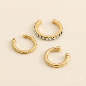 Puede incluir: Tres puños de oreja dorados. Uno está adornado con pequeños cristales transparentes. Otro tiene un diseño de cuerda trenzada, y el tercero es una banda lisa simple con extremos redondeados. Los puños de oreja se muestran sobre una superficie color crema.