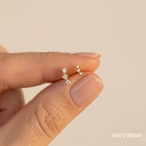 Puede incluir: Dos pendientes de oro, cada uno con múltiples gemas claras y brillantes. Un pendiente tiene tres piedras en disposición lineal, y el otro tiene tres piedras en disposición curva. Los pendientes se sostienen entre los dedos sobre un fondo neutro.