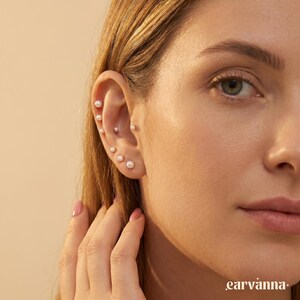 May include: A close-up of an ear displaying several small, round pearl earrings. The pearls are set in delicate, silver posts. The skin tone is fair, and the background is a soft, neutral colour. The brand name "earvanna" is visible.