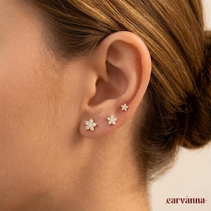May include: Three flower-shaped stud earrings with small, sparkling stones are displayed on an ear. The earrings are in graduated sizes, with the largest at the bottom and the smallest at the top. The ear is set against a blurred background.