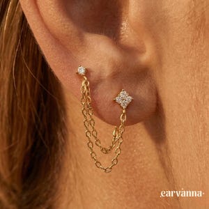 Könnte beinhalten: Goldene Doppelohrringe mit Ketten-Design. Ein Stecker hat einen einzelnen klaren Stein, während der andere eine quadratische Ansammlung klarer Steine hat. Die Ohrringe sind durch zwei zarte Goldketten verbunden.