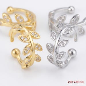 May include: Gold and silver ear cuffs. Each cuff has a leaf design with small, clear gemstones. The cuffs are designed to wrap around the ear. The image is set against a white background.