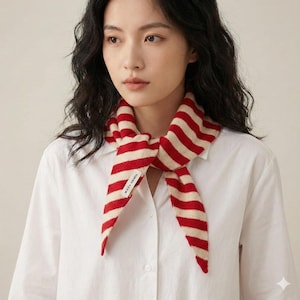 May include: A red and cream striped scarf tied around the neck. The scarf is made of a soft knit material and features bold, horizontal stripes. The scarf is worn over a white collared shirt. The scarf has a small tag with text.