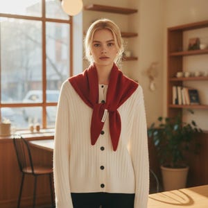 May include: A woman wearing a cream-colored ribbed cardigan with black buttons and a deep red scarf. The scarf is tied in a knot at the front. The background includes a window, a wooden table, and a bookshelf.