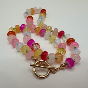 May include: A colorful beaded necklace featuring a variety of translucent and opaque beads in shades of pink, yellow, orange, and red. The necklace has a gold-tone toggle clasp. The beads are of various shapes and sizes.