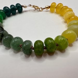 May include: A beaded bracelet featuring a gradient of green and yellow stones. The bracelet transitions from dark green to light green and then to yellow, with a gold-colored clasp. The beads are round and smooth, strung together with a delicate cord.