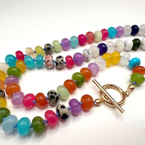 May include: A colorful beaded necklace featuring a variety of gemstone beads in shades of blue, green, yellow, orange, pink, purple, and white. The necklace has a gold-tone toggle clasp. The beads are approximately 0.5 inches in diameter.