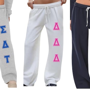 May include: Three pairs of sweatpants in gray, white, and navy blue. Each pair features a drawstring waist and a sorority or fraternity letter design in blue, pink, and green. The gray pants have blue letters, the white pants have pink letters, and the navy pants have green letters.