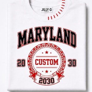 Op de afbeelding: Witte sweatshirt met het woord "MARYLAND" in zwart en rood. Een cirkelvormig ontwerp in rood en zwart toont "CUSTOM" en het jaar "2030". De halslijn heeft rode stiksels. Het merklabel "Jilly G" staat bovenaan.