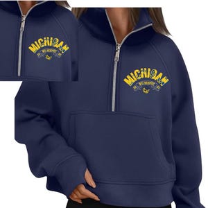 Puede incluir: Sudadera azul marino con cremallera de cuarto de cremallera y bolsillo de canguro. La sudadera presenta un logotipo de "Michigan Wolverines" amarillo y azul en el pecho. La cremallera es plateada.