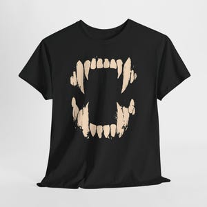 May include: Black t-shirt featuring a beige graphic of a set of stylized fangs. The design includes sharp, pointed teeth, creating a bold and edgy aesthetic. The shirt is made of a soft material.