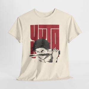 Puede incluir: Camiseta color crema con un gráfico de una persona sosteniendo una espada, con la palabra "YUTA" en letras rojas en la parte superior. La ilustración es en blanco y negro, creando un contraste llamativo con el tono neutro de la camiseta.