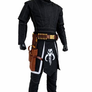 Puede incluir: Traje negro con costuras blancas, con una parte superior de cuello alto, pantalones y un cintur&oacute;n con detalles en forma de bala. Una funda marr&oacute;n y un gr&aacute;fico en blanco y negro en la falda completan el atuendo. El disfraz se exhibe en un maniqu&iacute;.