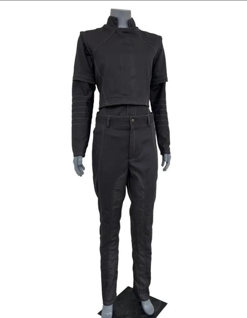 May include: Black two-piece outfit. The top is a cropped jacket with a high collar and long sleeves. The trousers are high-waisted with a button closure and a tailored fit. White stitching details the seams of the jacket and trousers.
