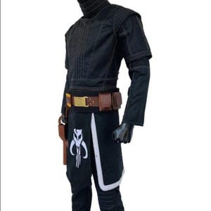 Puede incluir: Traje negro con cuello alto, mangas largas y cintur&oacute;n utilitario. El disfraz presenta detalles en blanco y un s&iacute;mbolo en la parte inferior de la pierna. Una funda est&aacute; sujeta al cintur&oacute;n.