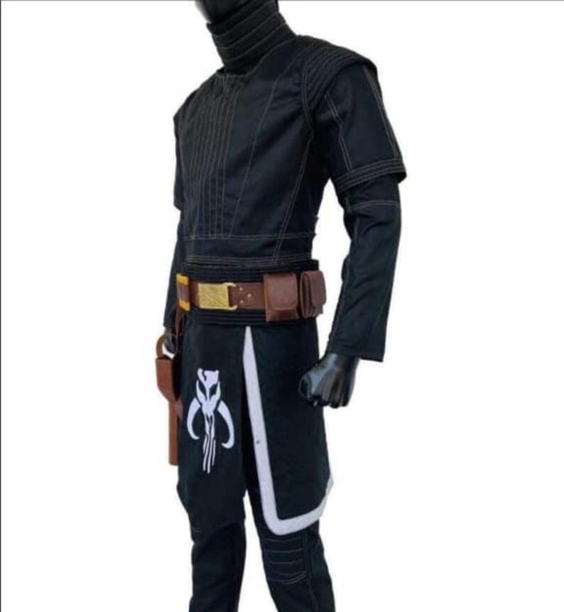 Puede incluir: Traje negro con cuello alto, mangas largas y cintur&oacute;n utilitario. El disfraz presenta detalles en blanco y un s&iacute;mbolo en la parte inferior de la pierna. Una funda est&aacute; sujeta al cintur&oacute;n.