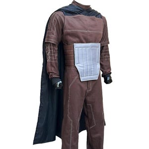 Puede incluir: Traje marr&oacute;n con capa negra. El traje tiene costuras blancas y una placa pectoral plateada. La capa est&aacute; drapeada sobre los hombros. El traje tiene mangas largas y pantalones. Los guantes son negros.