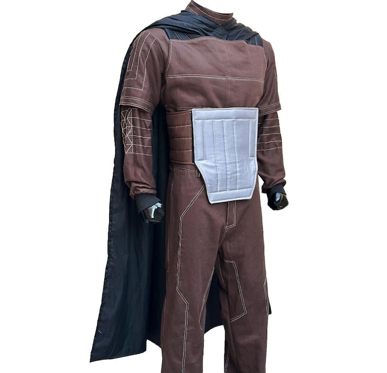 Puede incluir: Traje marr&oacute;n con capa negra. El traje tiene costuras blancas y una placa pectoral plateada. La capa est&aacute; drapeada sobre los hombros. El traje tiene mangas largas y pantalones. Los guantes son negros.