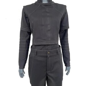 May include: A dark grey two-piece outfit. The cropped top has a high collar and long sleeves, and the matching trousers have a button closure. White stitching details are visible throughout the outfit, with horizontal stitching on the sleeves.