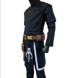 Puede incluir: Traje negro con cuello alto, mangas largas y cintur&oacute;n utilitario. El traje presenta detalles blancos y un s&iacute;mbolo en la parte inferior de la pierna. Una funda est&aacute; sujeta al cintur&oacute;n. El traje se exhibe en un maniqu&iacute;.