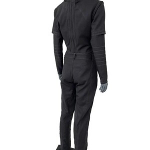 May include: Black jumpsuit with a high collar, long sleeves, and a fitted silhouette. The jumpsuit features visible stitching details and a belt at the waist. The sleeves have a layered design. The garment is displayed on a mannequin.