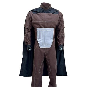 Puede incluir: Un disfraz marr&oacute;n con una capa negra. El disfraz tiene un cuello alto, mangas largas y costuras blancas. Un panel gris plateado est&aacute; en el pecho. El disfraz est&aacute; dise&ntilde;ado para parecerse a un personaje de una pel&iacute;cula de ciencia ficci&oacute;n.