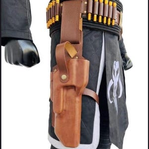 Puede incluir: Una funda de cuero marr&oacute;n con un cintur&oacute;n a juego, con una serie de objetos cil&iacute;ndricos con punta dorada. La funda est&aacute; sujeta a una prenda acolchada en blanco y negro. Un s&iacute;mbolo mandaloriano es visible en la prenda.