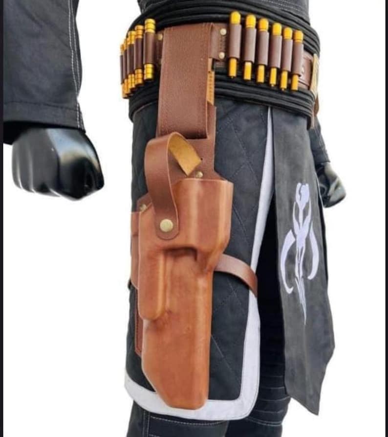 Puede incluir: Una funda de cuero marr&oacute;n con un cintur&oacute;n a juego, con una serie de objetos cil&iacute;ndricos con punta dorada. La funda est&aacute; sujeta a una prenda acolchada en blanco y negro. Un s&iacute;mbolo mandaloriano es visible en la prenda.