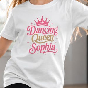 Könnte beinhalten: Ein junges Mädchen trägt ein weißes T-Shirt mit dem Schriftzug "Dancing Queen Sophia" in rosa und goldfarbenen Buchstaben, gekrönt von einer rosa Krone. Der Hintergrund enthält den Text "Dance is my superpower" und ein Neon-Schild mit der Aufschrift "Just Dance".