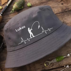 Könnte beinhalten: Grauer Fischerhut mit weißer Grafik eines Anglers, der einen Fisch einholt, integriert in eine Herzschlaglinie. Der Name "Lukas" ist über der Grafik aufgedruckt. Der Hut ist aus weichem Material.