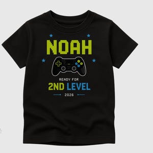 Könnte beinhalten: Schwarzes T-Shirt mit einem Videospiel-Controller-Motiv. Das Shirt zeigt den Namen "NOAH" in pixeligen grünen Buchstaben, darunter "READY FOR 2ND LEVEL 2026" in Blau.