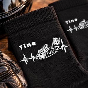 Könnte beinhalten: Ein Paar schwarze Socken mit einem weißen Grafikdruck eines Motorrads, einer Zielflagge und einer Herzschlaglinie. Der Text Tine ist in Weiß über der Motorradgrafik auf jeder Socke aufgedruckt, mit gerippten Bündchen am oberen Rand.