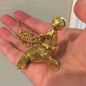 May include: A gold-tone necklace with a charm of a seal balancing a ball on its nose. The seal is detailed with fur and has a small collar.