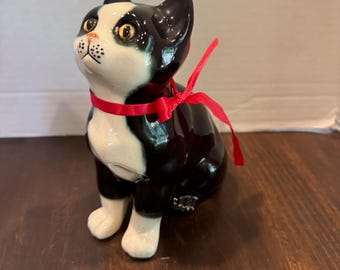 May include: A ceramic cat figurine with a black and white coat, a red ribbon around its neck, and golden eyes. The cat is in a sitting position, looking upwards. The figurine has a glossy finish and is placed on a wooden surface.