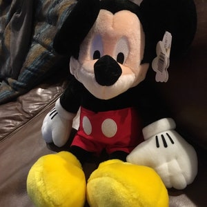 May include: A plush Mickey Mouse toy with black ears, a black nose, and yellow shoes. He is wearing red shorts with white polka dots. The toy is sitting on a brown leather sofa.