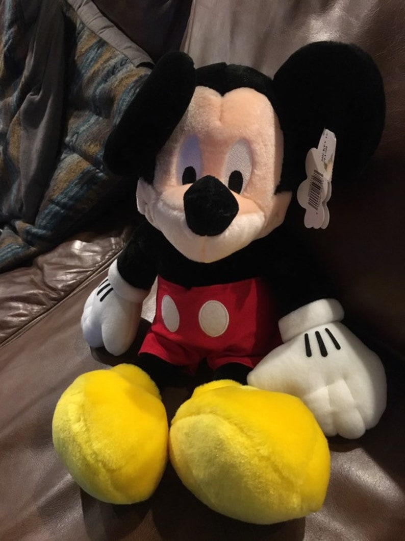 May include: A plush Mickey Mouse toy with black ears, a black nose, and yellow shoes. He is wearing red shorts with white polka dots. The toy is sitting on a brown leather sofa.