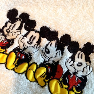 Puede incluir: Toalla blanca con cuatro personajes de Mickey Mouse bordados. Cada Mickey Mouse lleva pantalones cortos rojos y tiene ojos negros y zapatos amarillos.