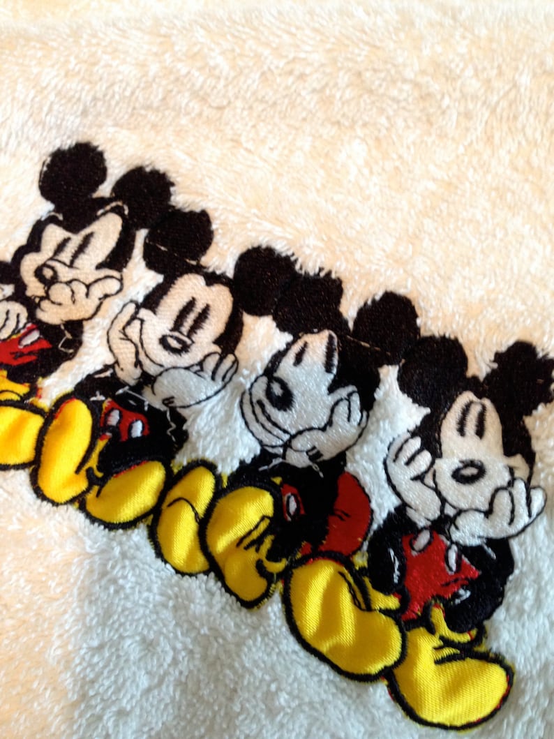 Puede incluir: Toalla blanca con cuatro personajes de Mickey Mouse bordados. Cada Mickey Mouse lleva pantalones cortos rojos y tiene ojos negros y zapatos amarillos.